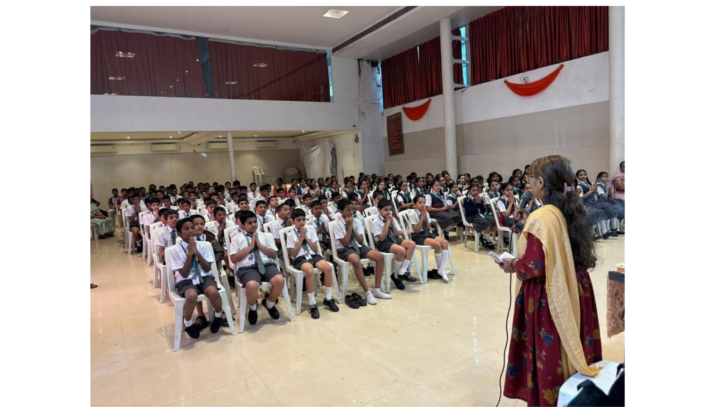 विद्यालय के सभागार में सैकड़ों विद्यार्थी हाथ जोड़े श्लोक दोहराते हुए, सामने माइक लिए गीता परिवार की प्रशिक्षिका।